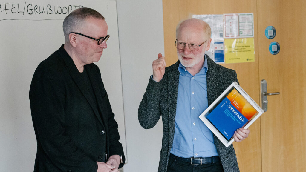 Dr. Olaf Gaus (l.) übergibt Prof. Dr. rer. nat. Rainer Brück symbolisch das Titelblatt des demnächst im Springer-Verlag erscheinenden Buchs „Datenmedizin“, das Prof. Brück gewidmet ist.